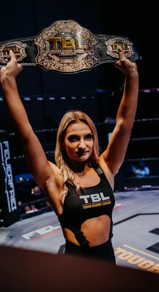 TBL Events ring girl holding a championship belt above her head inside an MMA fight cage during a combat sports event.